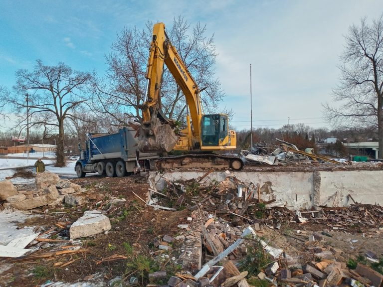 Excavator clearing a demolished building beside a dump truck